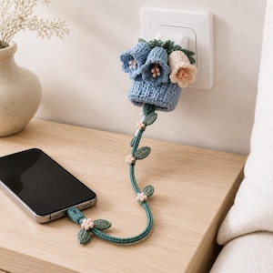 以下が含まれることがあります： 壁の充電器にフィットする青いベースを備えたかぎ針編みのケーブルプロテクター。青とクリーム色のベル型の花と緑の葉で装飾されています。ベースから伸びる長く細い緑色のコードには、小さなピンクの花と葉が飾られています。