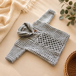 May include: A light gray long-sleeve pullover sweater featuring a textured bobble stitch pattern, a shawl collar with a single brown wooden button, and a diamond-shaped openwork lattice design on the front panel, crafted from yarn.