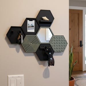 May include: A modular wall-mounted organizer system composed of interconnected black and sage green hexagonal units. The set includes a mirror, a small shelf with a seashell, a photo clip, hooks for keys and sunglasses, and textured panels.
