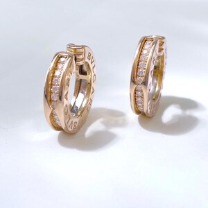 May include: A pair of gold hoop earrings featuring a central row of small clear round-cut diamonds. The outer edges of the hoops are engraved with the text BVLGARI in capital letters. The earrings have a polished metallic finish and a hinged design.