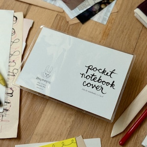 May include: A clear PVC pocket notebook cover with a white insert featuring black text that reads pocket notebook cover, fits B7 notebooks 3.5x4.9, and stray margins with a rabbit logo. The insert also lists size 7.5x5.3, material PVC, and made in USA.