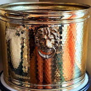 May include: A cylindrical brass planter featuring a hammered texture across its exterior surface. The vessel is adorned with a prominent three-dimensional lion head ornament that holds a circular ring in its mouth, centered on the front side.
