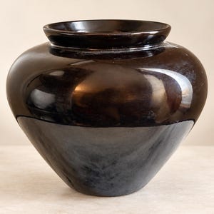 Puede incluir: Un jarrón de cerámica de color marrón oscuro brillante con un cuerpo ancho y redondeado que se estrecha hacia una base pequeña. El recipiente presenta una superficie lisa y reflectante con un borde circular superior ligeramente abierto.