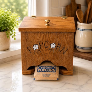May include: A rectangular wooden popcorn dispenser with a hinged lid, a brass knob, and a front cutout. The word POPCORN is printed on the front with small cloud graphics. A single brown paper microwave popcorn bag is visible in the dispenser opening.