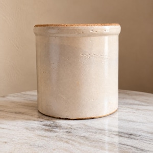 May include: A cylindrical ceramic crock with a smooth, off-white glazed finish and a flat, textured cork lid. The vessel features a slightly flared rim and a sturdy, straight-sided body, showcasing subtle surface variations in the glaze.