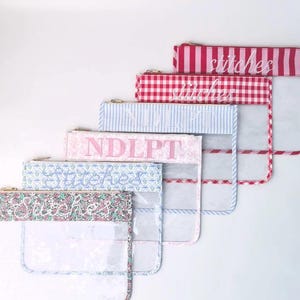 Può includere: Un set di sei bustine rettangolari trasparenti con chiusura a zip. Ogni bustina presenta un bordo superiore in tessuto unico con motivi che includono righe rosse, quadretti vichy rossi, righe blu, fiori rosa, fiori blu e stampe floreali.