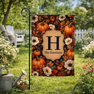 May include: A rectangular garden flag featuring a dark background with orange pumpkins, white flowers, and autumn leaves. A central tan scalloped label displays the letter H and the text The Harrison in dark blue, hanging on a black metal stand.