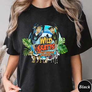 May include: A black t-shirt featuring a graphic print of two cartoon characters, a cheetah, a monkey, and a zebra. The text "WILD KRATTS" is displayed in large orange letters above the words "AMAZIN' AMAZON ADVENTURE" in a blue circle.