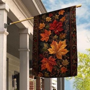 May include: A rectangular garden flag hanging from a gold-colored metal pole. The flag features a dark background with a pattern of vibrant autumn maple leaves in shades of red, orange, and yellow, framed by ornate, dark decorative borders.
