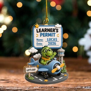 May include: A hanging Christmas ornament featuring a green dinosaur wearing glasses in a blue airplane on a runway. A sign reads LEARNER'S PERMIT, Name: LUCAS, Date: 2026. A small sign says LET'S GO! The design includes a control tower, clouds, and stars.
