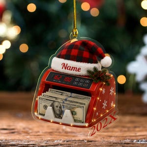 May include: A red money counting machine ornament featuring a stack of one hundred dollar bills, a red and black buffalo plaid Santa hat with the text Name, and the year 202X printed in red. The ornament hangs from a gold string.