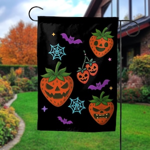 May include: A black garden flag featuring a pattern of orange strawberry jack-o'-lanterns with green stems, light blue spiderwebs, purple bats, and small colorful stars. The strawberries display various carved facial expressions.