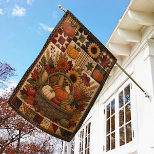 May include: A rectangular outdoor flag hanging from a metal pole, featuring a printed quilt-style design of a woven basket filled with pumpkins, sunflowers, wheat, and autumn leaves, accented by geometric star patterns in orange, brown, and gold.