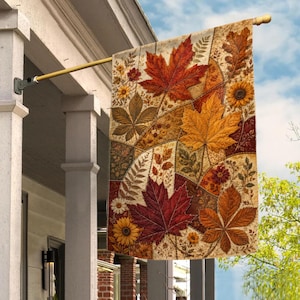 May include: A rectangular garden flag hanging from a gold-colored metal pole, featuring a patchwork design of autumn leaves in shades of red, orange, yellow, and brown, interspersed with small floral patterns and various botanical leaf silhouettes.