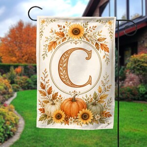 May include: A rectangular garden flag featuring a large, ornate letter C centered within a circular frame. The design is decorated with autumn-themed illustrations including sunflowers, orange and green pumpkins, and fall foliage on a cream background.
