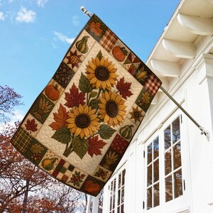 May include: A rectangular garden flag featuring a quilted-style print of sunflowers, orange pumpkins, and red maple leaves. The border consists of patchwork squares with patterns of plaid, wheat, acorns, and berries on a cream-colored background.