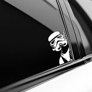 May include: A white vinyl decal of a Stormtrooper helmet peeking out from the side, applied to a dark car window. The graphic features bold black outlines and solid white fill, depicting the iconic character looking around a corner edge.