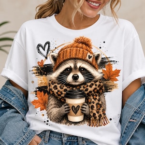 May include: A white t-shirt featuring a graphic of a raccoon wearing an orange knit beanie and a leopard print scarf. The raccoon holds a coffee cup with a heart design. The background includes orange maple leaves, black brush strokes, and hearts.