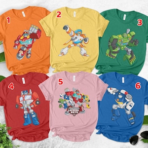 May include: Six short-sleeved t-shirts in orange, yellow, green, red, light pink, and blue. Each shirt features a graphic of a Transformers Rescue Bots character, with the pink shirt displaying a group illustration and the text Transformers Rescue Bots.