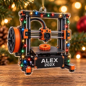 May include: A clear acrylic Christmas ornament shaped like a 3D printer with orange and black accents. The printer features a spool of orange filament, a printed orange gear on the build plate, and the text ALEX 202X. Multi-colored fairy lights wrap around the frame.