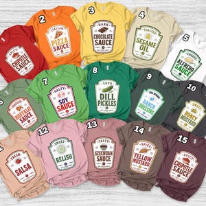 May include: A collection of fifteen short-sleeved t-shirts in various colors, each featuring a graphic label design resembling a condiment bottle. The labels include text such as Chipotle Sauce, Pizza Sauce, Chocolate Sauce, Sesame Oil, Alfredo Sauce, Buffalo Sauce, Soy Sauce, Dill Pickles, Ranch Dressing, Honey Mustard, Salsa, Relish, Szechuan Sauce, Yellow Mustard, and Chipotle Sauce.