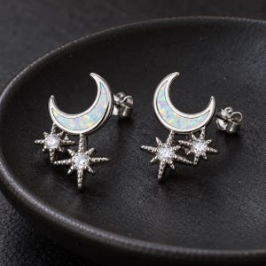 Pode incluir: Um par de brincos prateados em forma de meia-lua incrustados com material branco iridescente, acentuados por dois pingentes em forma de estrela com pedras redondas transparentes e brilhantes e fechos de pino.