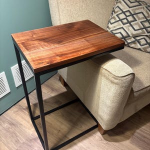 Puede incluir: Una mesa auxiliar en forma de C con un tablero rectangular de madera marrón oscura con veta visible. La mesa está sostenida por una estructura de metal negro delgada que forma una base rectangular continua y un soporte vertical.