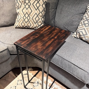 Puede incluir: Una mesa auxiliar rectangular de color marrón oscuro con estructura metálica. La encimera tiene un diseño de vetas de madera. La mesa está colocada junto a un sofá gris con cojines estampados. Las dimensiones de la mesa son de unos 61 cm de largo, 30 cm de ancho y 61 cm de alto.