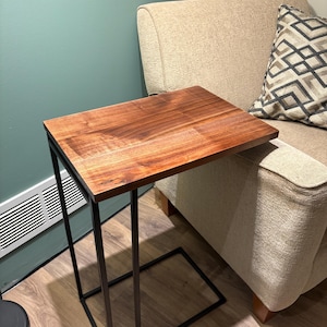 Puede incluir: Una mesa auxiliar rectangular con una tapa de madera oscura y un marco de metal negro. La mesa está diseñada para deslizarse debajo de un sofá o una silla. La tapa de madera tiene un color marrón cálido y el marco de metal tiene un diseño minimalista.