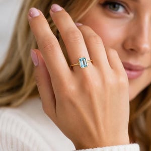 May include: A gold ring featuring a single emerald-cut light blue gemstone set in a bezel setting. The band is thin and polished, showcasing a clean, simple design that highlights the rectangular shape and clarity of the central blue stone.