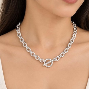 May include: A silver-toned chain necklace featuring thick, rounded oval links and a prominent toggle clasp closure at the center. The necklace rests around the neck, showcasing a polished metallic finish and a classic, uniform link design throughout.