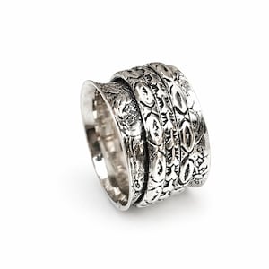 Puede incluir: Un anillo giratorio plateado con intrincados patrones florales y geométricos. El anillo presenta una banda ancha con múltiples secciones giratorias, creando un diseño texturizado y detallado. El anillo está sobre un fondo blanco.