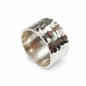 Puede incluir: Un anillo ancho de plata con textura martillada. El anillo tiene una banda gruesa y una superficie brillante y reflectante. El diseño martillado crea una apariencia texturizada única. El anillo está sobre un fondo blanco.