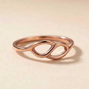 Peut inclure: Une bague en or rose composée d'un anneau lisse et continu qui forme deux ouvertures interconnectées en forme d'amande au centre. Le métal présente une finition polie et réfléchissante, avec un design symétrique créant un aspect ajouré fluide.
