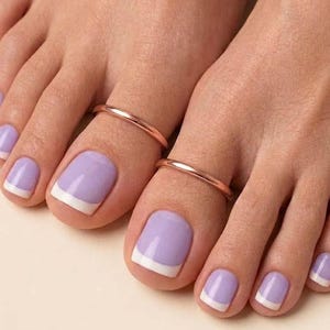 Peut inclure: Deux anneaux d'orteil en or rose lisse et poli portés sur le deuxième orteil de chaque pied. Les ongles sont vernis d'une teinte lavande claire avec une manucure française blanche. Le texte Kanhajewellers1 est visible en bas à droite.