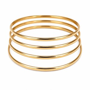 Pode incluir: Um conjunto de quatro pulseiras rígidas douradas empilhadas. Cada pulseira apresenta uma superfície lisa, polida e arredondada com uma forma circular consistente. As pulseiras estão dispostas verticalmente, criando uma aparência em camadas com superfícies reflexivas.