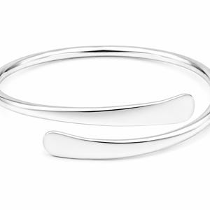 Könnte beinhalten: Ein silberfarbenes, offenes Armreif-Armband. Das Armband hat eine glatte, polierte Oberfläche und ein minimalistisches Design. Die Enden des Armbands sind leicht abgeflacht und gebogen, was einen modernen und eleganten Look erzeugt.