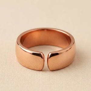 Puede incluir: Un anillo de tono oro rosa pulido con una banda ancha y lisa de diseño abierto. La superficie metálica es reflectante y presenta bordes redondeados, creando un aspecto limpio y continuo que se curva para formar una pequeña abertura al frente.