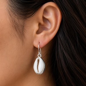 Peut inclure: Une boucle d'oreille pendante en argent avec un coquillage cauri blanc enchâssé dans une monture en argent lisse. La boucle d'oreille est suspendue à un crochet simple en argent, mettant en valeur la forme ovale naturelle et la fente centrale.