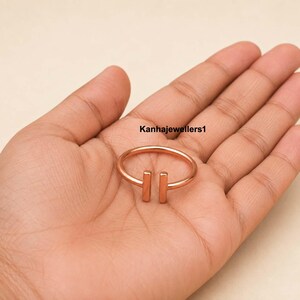 Può includere: Un anello aperto color oro rosa con due estremità a barra verticale che si fronteggiano con un piccolo spazio tra loro. La superficie metallica è liscia e lucida, riflettendo la luce. Il testo Kanhajewellers1 è visibile sopra l'anello.
