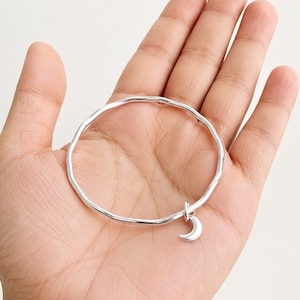 Puede incluir: Una pulsera tipo brazalete de color plateado con una textura ondulada e irregular a lo largo de la banda. La pulsera presenta un pequeño dije colgante en forma de media luna unido a un lado. El metal tiene un acabado pulido y brillante.