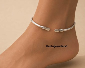 Puede incluir: Una tobillera abierta de tono plateado con una banda lisa y pulida que presenta tres anillos decorativos envueltos en alambre cerca de cada extremo cónico. La pieza metálica rodea el tobillo y el texto Kanhajewellers1 es visible en la piel.