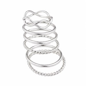 Pode incluir: Uma pilha de anéis de prata em vários designs. Os anéis incluem um estilo de corda torcida, uma faixa lisa e um design de símbolo de infinito. Os anéis são exibidos em um fundo branco.