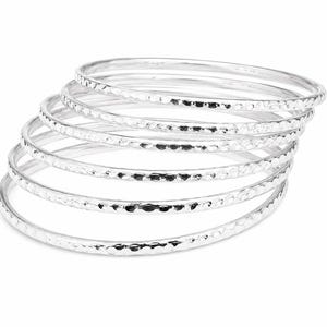 Può includere: Una pila di sette braccialetti d'argento con una texture martellata. I braccialetti sono rotondi e hanno una superficie lucida e riflettente. I gioielli sono presentati su uno sfondo bianco.