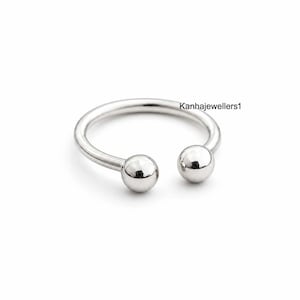 Puede incluir: Un anillo en forma de herradura de color plateado pulido con dos extremos esféricos lisos. La superficie metálica es reflectante. El texto Kanhajewellers1 está impreso en negro cerca del lado derecho de la banda circular abierta.