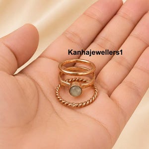 Peut inclure: Un ensemble de trois bagues empilées de couleur or rose. La bague supérieure est un anneau lisse, la bague centrale présente une pierre ronde vert pâle, et la bague inférieure a une texture torsadée. Le texte Kanhajewellers1 est visible.
