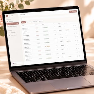 Könnte beinhalten: Ein silberner Laptop, der eine digitale Gästelisten-Oberfläche auf dem Bildschirm anzeigt. Die Software zeigt eine Tabelle mit Spalten für Namen, Status und Tischnummern. Die Kopfzeile lautet All Guests und enthält ein Seitenmenü.
