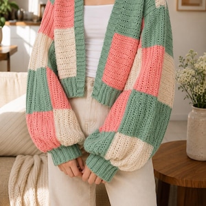 Pode incluir: Um cardigã de crochê de manga comprida com um padrão de blocos de cores em verde sálvia, rosa suave e creme. A peça tem decote em V, punhos canelados e bainha canelada, usada sobre uma blusa branca e calças de ganga claras.