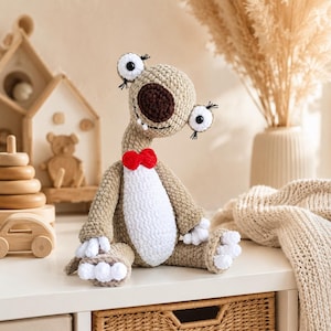 Peut inclure: Une peluche crochetée avec un corps beige, un ventre blanc et un nez marron foncé. La créature possède deux grands yeux blancs avec des pupilles noires et de longs cils, un petit nœud papillon rouge au cou et des détails blancs sur les mains et les pieds.