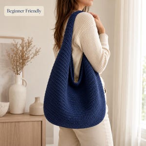 Peut inclure: Un sac hobo en crochet bleu marine profond avec un motif de mailles côtelées texturées et une large bandoulière intégrée. Le sac présente une silhouette souple et déstructurée, avec une petite étiquette rectangulaire indiquant Beginner Friendly.